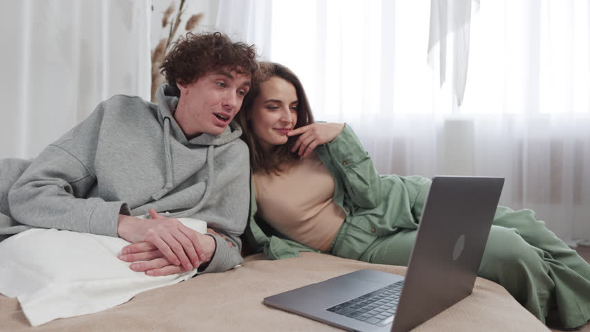 Young couple smiles and chats casually as they browse on a laptop, sharing a spontaneous moment of togetherness in their minimalist bedroom. Slow motion. Royalty-Free Stock Footage #3479015241