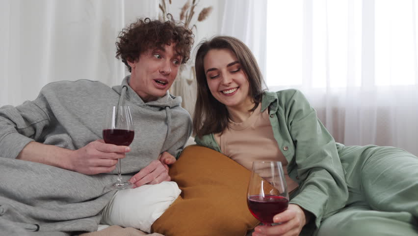 Comfortably lounging, a couple enjoys a light-hearted moment, wine glasses in hand, surrounded by a chic, minimalist decor. Their joy is infectious. Slow motion. 4K Royalty-Free Stock Footage #3479017745
