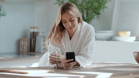 Video of smiling young woman enjoying a cup of coffee while using her mobile phone in the living room at home.