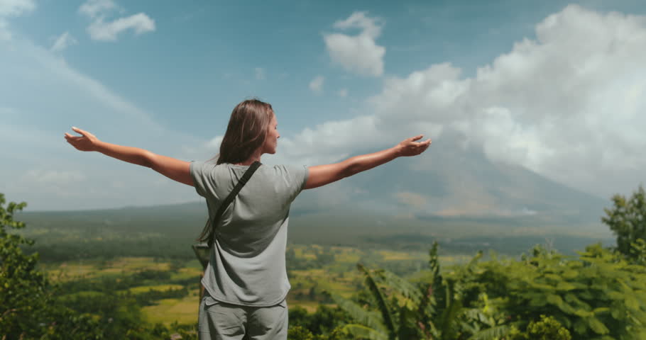 A woman standing a top a vibrant green hillside overlooking a scenic landscape. The hillside is rich with vegetation under the blue skies.