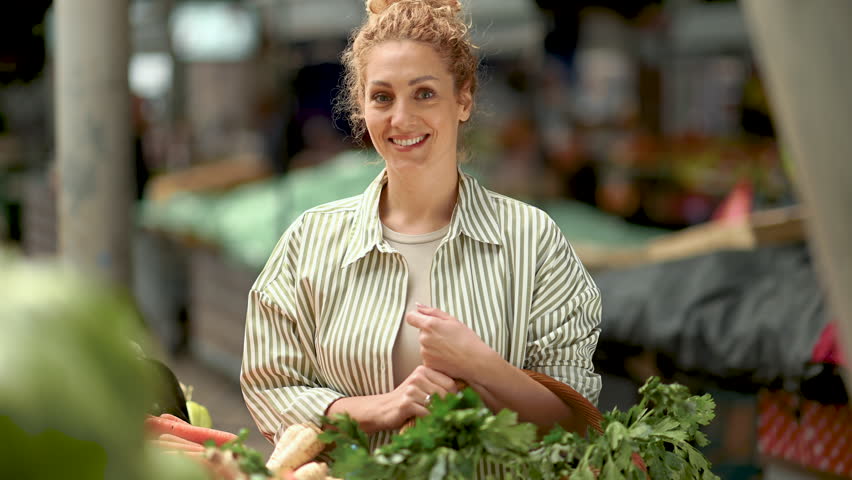 Portrait of a cheerful customer with healthy habits standing with wicker shopping basket at green market and smiling at the camera. Smiling female shopper near market stall posing with basket in hands