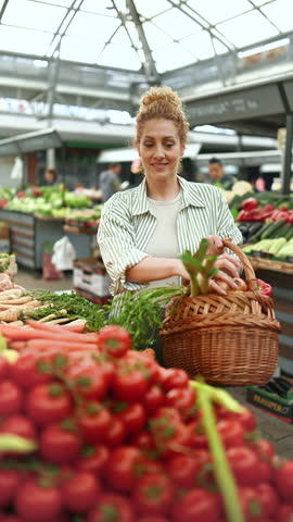 Smiling healthy shopper woman with wicker shopping basket in hands choosing fresh tomato at farmers market. Female customer with basket purchasing organic groceries at greengrocery market.