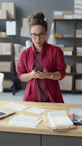 Vertical shot of female manager speaking on phone, using laptop and checking correspondence at work in delivery service office