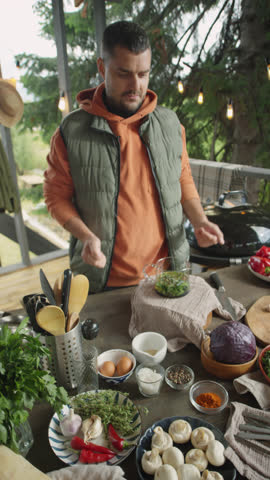 Vertical shot of food blogger standing at table with ingredients on outdoor terrace and telling recipe on camera while filming video recipe
