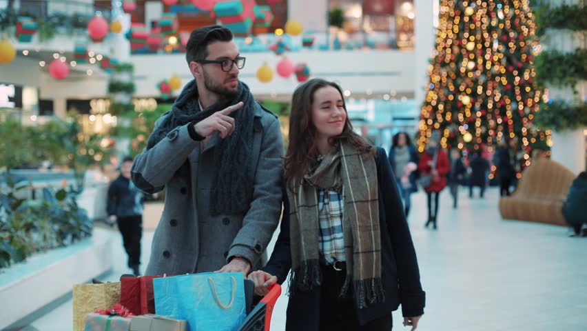 Close up lovely couple with Christmas present walk in a mall smiling feel happy shopper family woman tree christmas shopping man shop store portrait gift holiday shopping slow motion