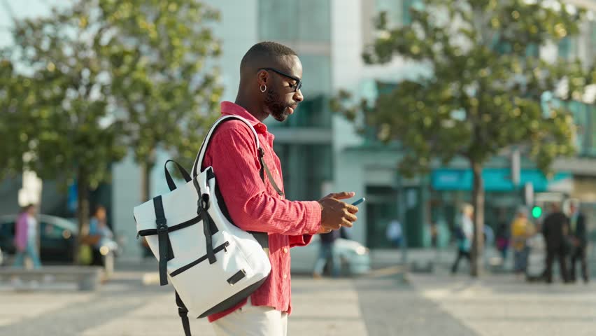 Cool young African Black man hipster student or tourist wearing sunglasses and backpack walking on city street outdoors holding smartphone using mobile cell phone technology device.