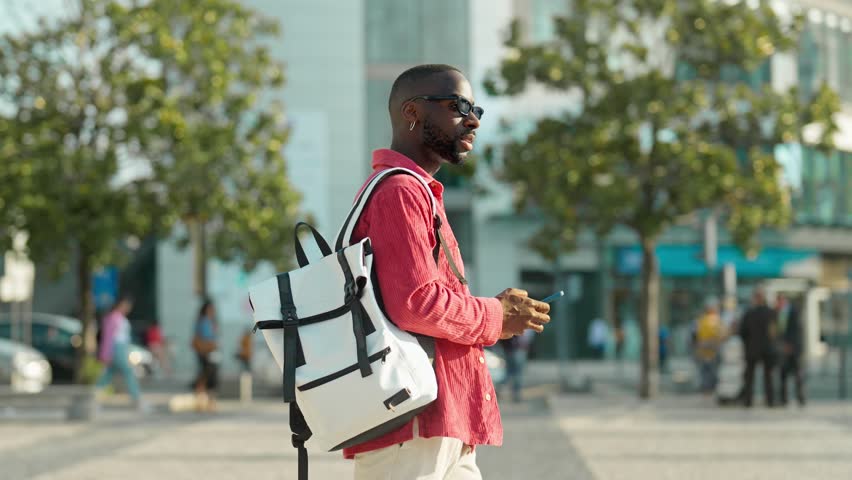 Cool young African Black man hipster student or tourist wearing sunglasses and backpack walking on city street outdoors holding smartphone using mobile cell phone technology device.