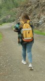 Vertical video. Following a Young Female Traveler, She Walks Along a Rocky Mountain Road Amidst Trees. Bright Orange Backpack on her Back.