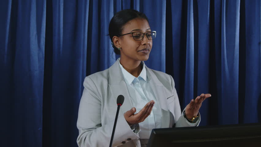 Young female politician standing at lectern with microphone and giving speech in front of public and journalists during press conference