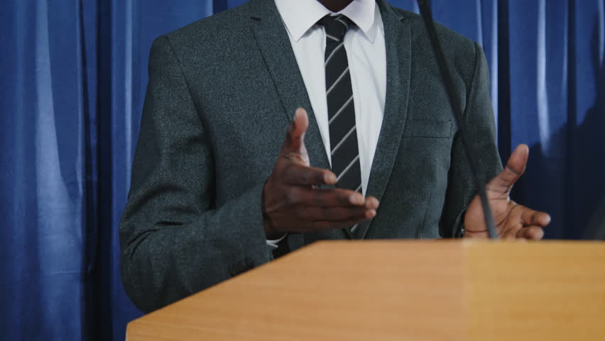 Tilt up shot of young Black politician standing at lectern with microphone and giving speech during debates or press conference