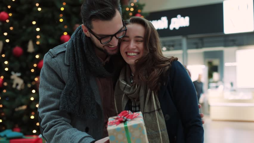 Close up young man hugging girlfriends and gives Christmas gift in the mall background Christmas tree couple shopping family woman love relationship romantic smiling celebrating slow motion