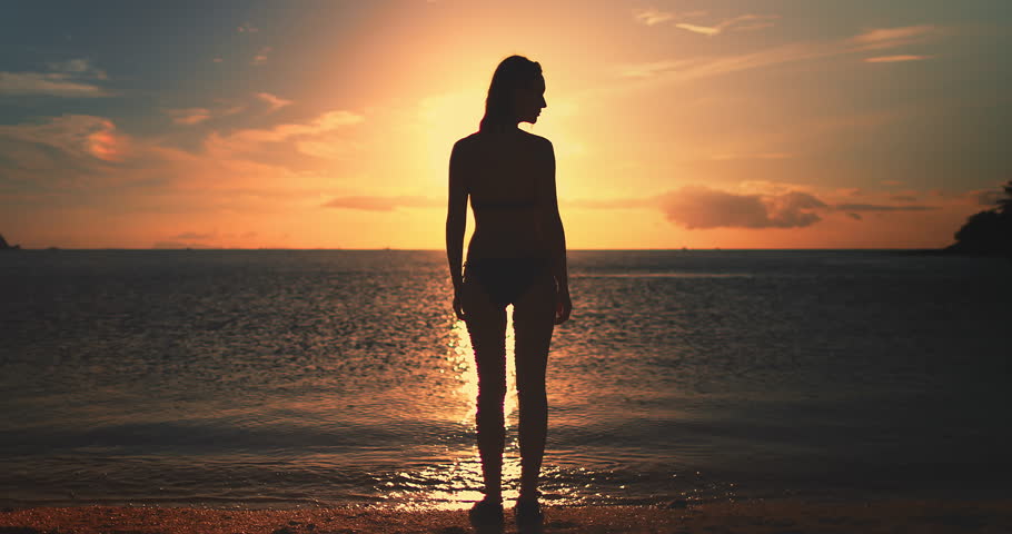 Woman stands on Koh Ma Beach at Koh Phangan Island in Thailand during sunset, with the sky painted in warm hues and gentle waves breaking on the shore.