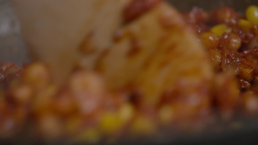 Corn and chickpeas, mixed with red spicy seasoning, are combined with a wooden spoon in a traditional Asian dish. Extreme close-up. Slow motion. 4k