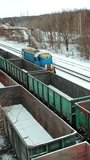 A freight train is passing by rail along rows of containers outside the city in the station area in the winter. Aerial view. Vertical video
