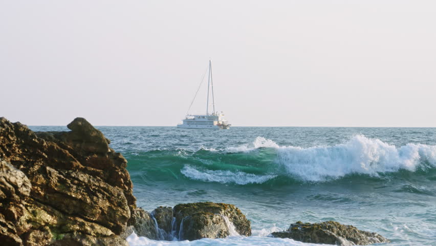 Sailboat navigates through ocean waves near rocky coastline. Seafaring yacht on blue sea adventure. Maritime travel, leisure sailing. Nautical vessel on water with rocky shore in foreground. Slowmo