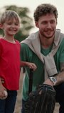 Vertical video: A dad with curly hair with stubble in a Green T-shirt with a baseball glove on his hand and near his little blond son in a red T-shirt with a baseball bat pose together on the