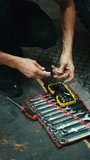 Vertical video: close-up: a male mechanic with his sinewy hands selects the tool he needs among a set of tools on the floor in the workshop
