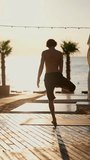 Vertical video: a guy stands on one leg and meditates with his hands up on a sunny beach