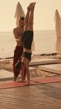 Vertical video: a brunette guy in red pants with a bare torso helps a girl in a black sports summer uniform to stand on her hands with her head down on a sunny beach on a red carpet