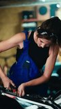 Vertical video: A tired brunette mechanic girl in a black top and blue overalls finishes repairing a bike in her garage workshop. The end of a hard working day for a mechanic girl in a workshop garage