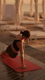 Vertical video: Rear view of a brunette girl in a black sports summer uniform doing a plank on one arm and reaching up with the other. Yoga and sports class on a sunny beach with umbrellas in summer