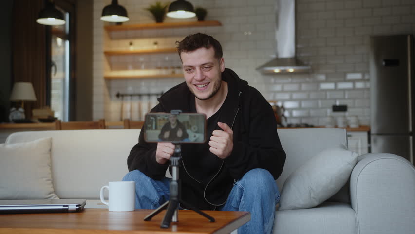 Young influencer vlogger man sit on the couch recording video blog on mobile camera tripod talk for personal media channel, streaming live session blogging broadcast at home.