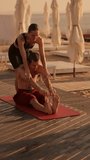Vertical video: a brunette girl in a black top helps a brunette guy do stretches and reach for his feet, sitting on a red mat on the beach in the summer
