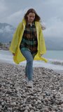 Vertical video. Young Woman in a Yellow Raincoat Strolling along the Seaside Beach in a Storm, with Mountains and Dark Clouds in the Background. In Slow Motion.