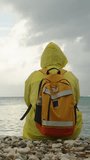 Vertical video. Overcast weather, clouds over the mountains and the sea, a woman in a yellow raincoat sadly sits on the beach and gazes into the distance. Circular motion.