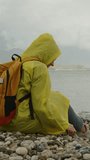 Vertical video. A woman in a yellow raincoat with a backpack sits and throws a stone into the sea. Rainy weather and overcast sky. Loneliness and sadness.