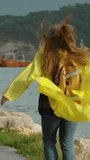 Vertical video. With arms outstretched to the wind, a woman in a yellow raincoat walks along the seaside promenade in stormy weather. Her long hair and raincoat billow in the wind. Mountains and a shi