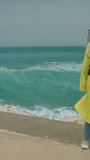 Vertical video. A high wave approaches the woman in a yellow raincoat with a backpack. Crashing against the rocks, it shatters into tall splashes, covering everything around. The woman narrowly avoids