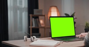 Man using laptop with chroma key screen. Male sitting at table and looking at green mock up of laptop. Man working at modern living room. Male looking at green template laptop display.
