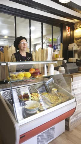 A senior woman with a warm smile serves a variety of fresh deli items from behind the glass counter at a local takeaway. Royalty-Free Stock Footage #3479716633