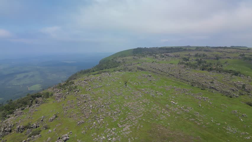 A high aerial rotation looking down onto grassy, rocky terrain, clusters of indigenous trees, a deep ravine embedded with green foliage, and a hazy valley under a blue, cloudy sky in 4K.