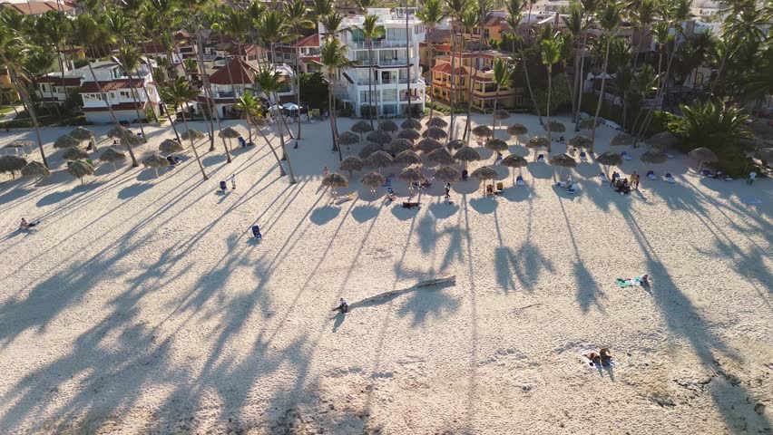 Panoramic view from above captures the beach at sunset, adorned with sun loungers, umbrellas, and palm trees, while tourists delight in their tropical island getaway. Dominican Republic, Punta Cana