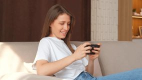 Footage of young charming woman playing online games on smartphone while sitting on sofa