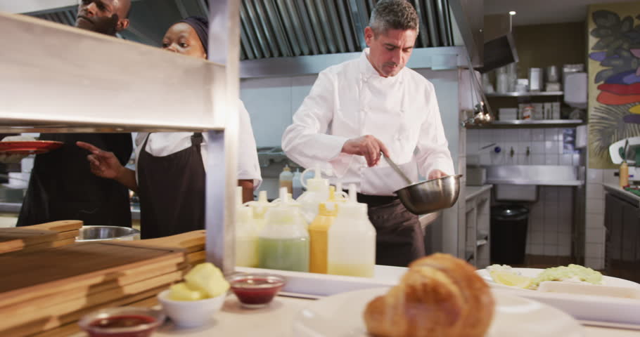 Front view of a Caucasian male chef working in a busy restaurant kitchen, garnishing plates of food ready to be served, female and male cooks working in the background