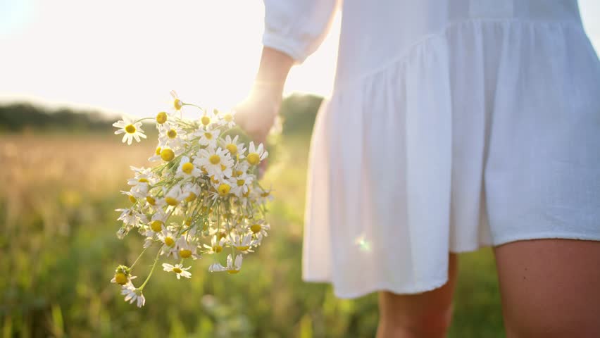 Woman in white dress walk along rural road enjoying nature, legs close-up. Female positive romantic holding bouquet of wildflowers in hands. Resting in village, freedom, female relax concept.