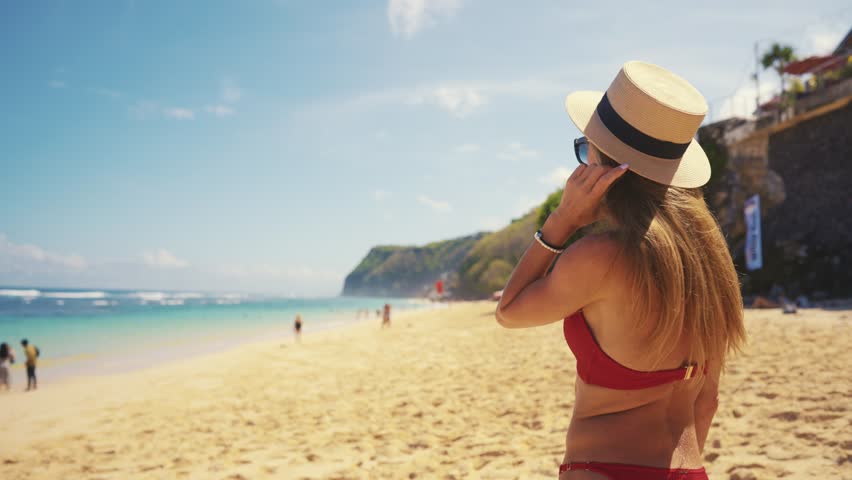 Blonde woman in red bikini hat admire on ocean beach in tropical paradise island with turquoise water waves, white sand in sunny weather. Girl admire seascape. Beach resting, travel, tourism.