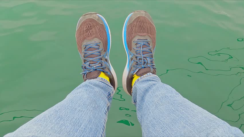 Man dangling his feet into the water. View of legs in jeans and sneakers dangling above the surface of the sea. Close-up.