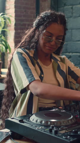 Vertical medium portrait of stylish young African American woman wearing casual outfit playing music using DJ mixer in loft studio