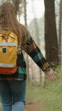 Vertical video. A Rear View of a Young Woman Hiking with a Backpack, Traveling Alone Through the Forest, Touching the Tall Grass. Selective Focus.