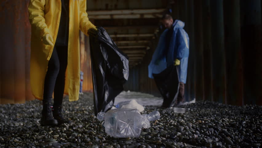 Man and woman in yellow and blue raincoat are cleaning up on the coast. Girl and guy collect garbage and waste on a stone beach of the sea or ocean. Female and male take care of planet and environment Royalty-Free Stock Footage #3479927289