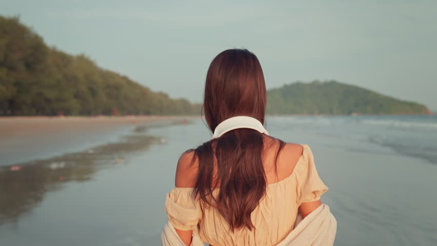 Relax asian woman wearing headphones walking in the sea beach at golden sunset - Female tourist listening to music on summer vacation