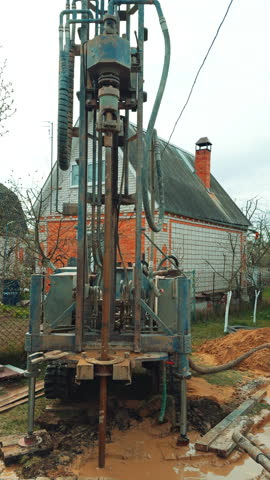 Boring the well with the help of special machinery. Drilling equipment producing the water bore. Private cottage at backdrop. Vertical video.