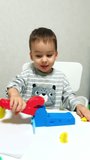 Busy little Caucasian boy sitting a desk plays with device molding plasticine. Happy kid sculpturing from clay at home. Vertical video.