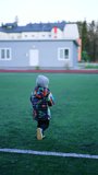Little baby boy in warm clothes runs to the stadium. Toddler kid plays football kicking the ball. Vertical video.