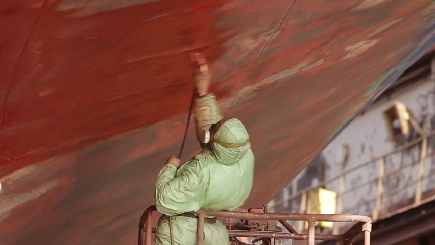 Worker in protective gear applies red paint to ship hull in dry dock. Industrial painting, maintenance against rust, maritime vessel refurbishment process. Shipyard worker, safety equipment in use.