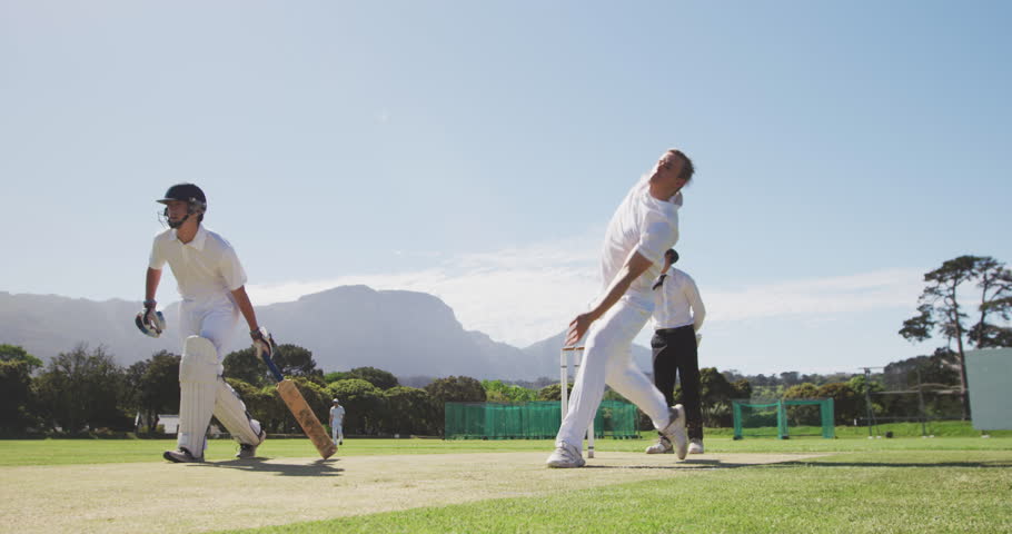 Front view of a teenage Caucasian male cricket player wearing whites, bowling the ball on the pitch during a cricket match, with an umpire standing in the background and a batsman running.